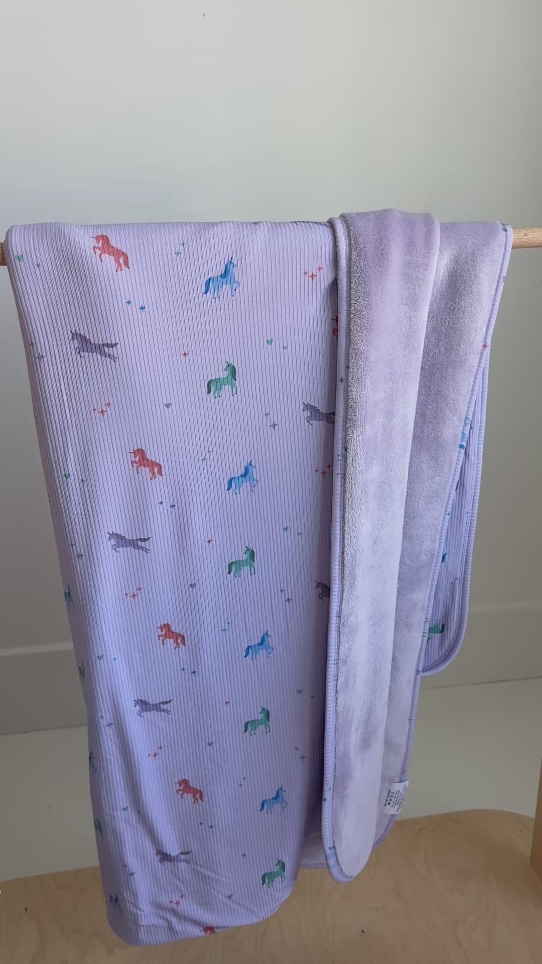 Light purple baby blanket with colorful unicorn patterns hanging on a rack