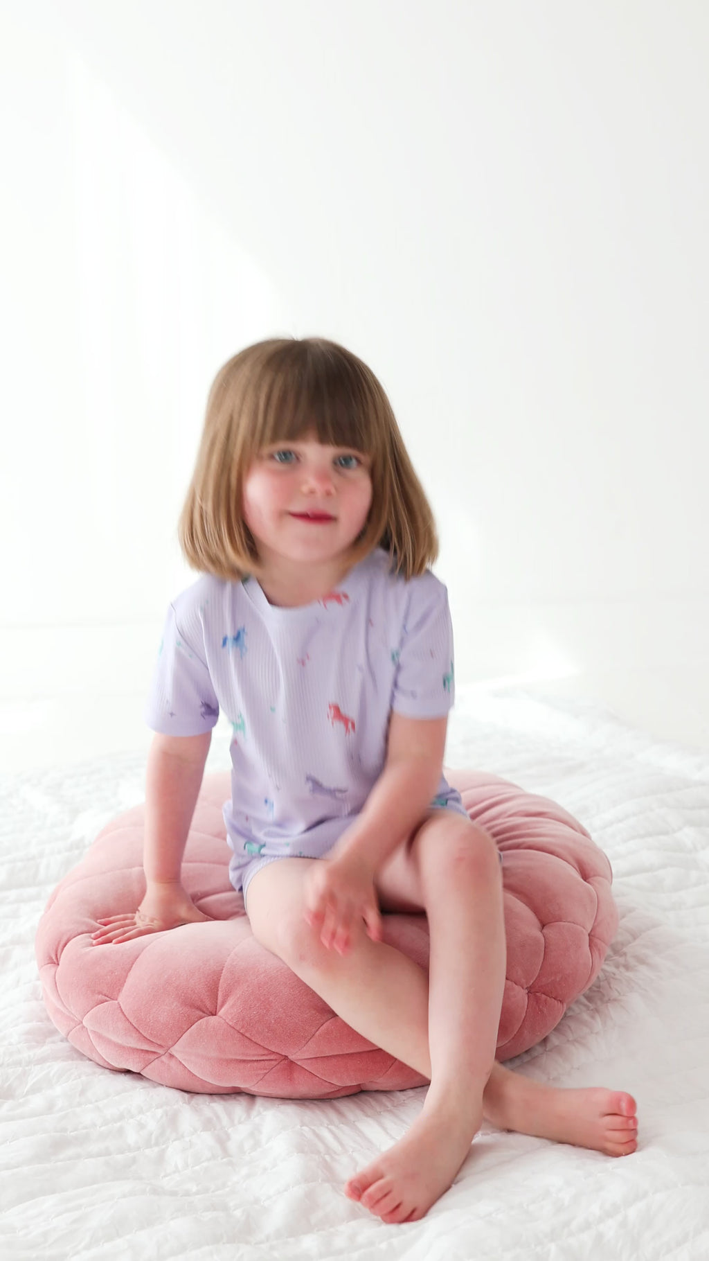 Video of a young girl wearing a light purple pajama shorts set with unicorn patterns, sitting on a pink cushion with a white background