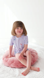 Video of a young girl wearing a light purple pajama shorts set with unicorn patterns, sitting on a pink cushion with a white background
