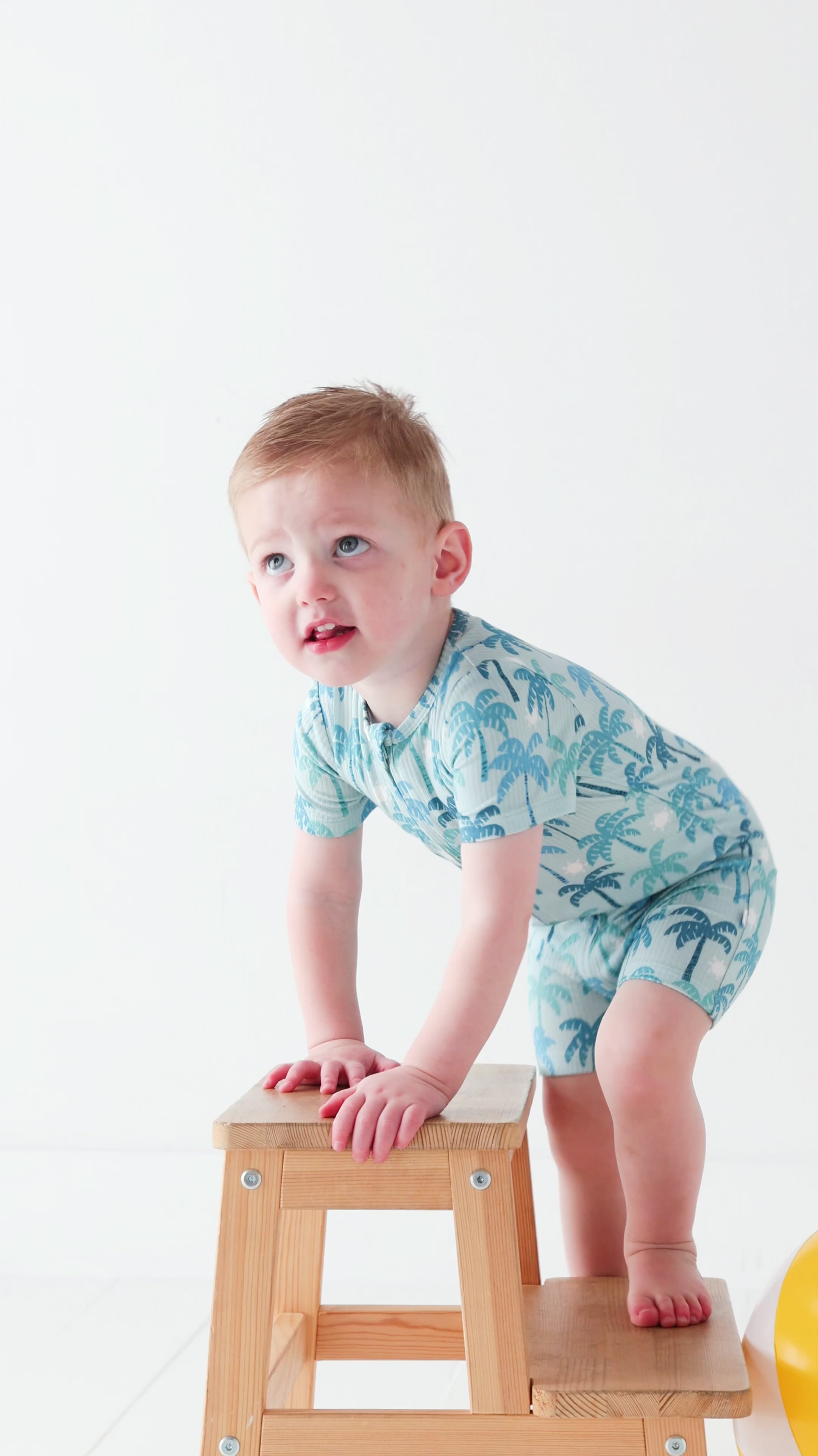 Child wearing a blue palm tree patterned onesie on a wooden stool against a white background