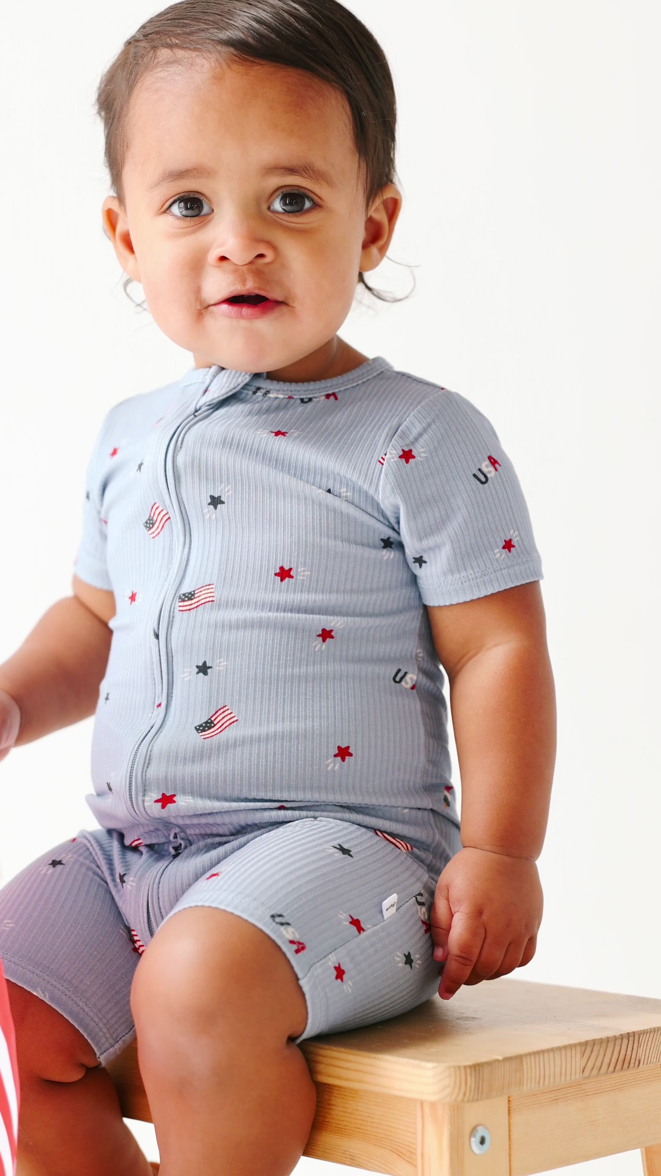 Child wearing a short romper with American-themed pattern sitting on a wooden stool.