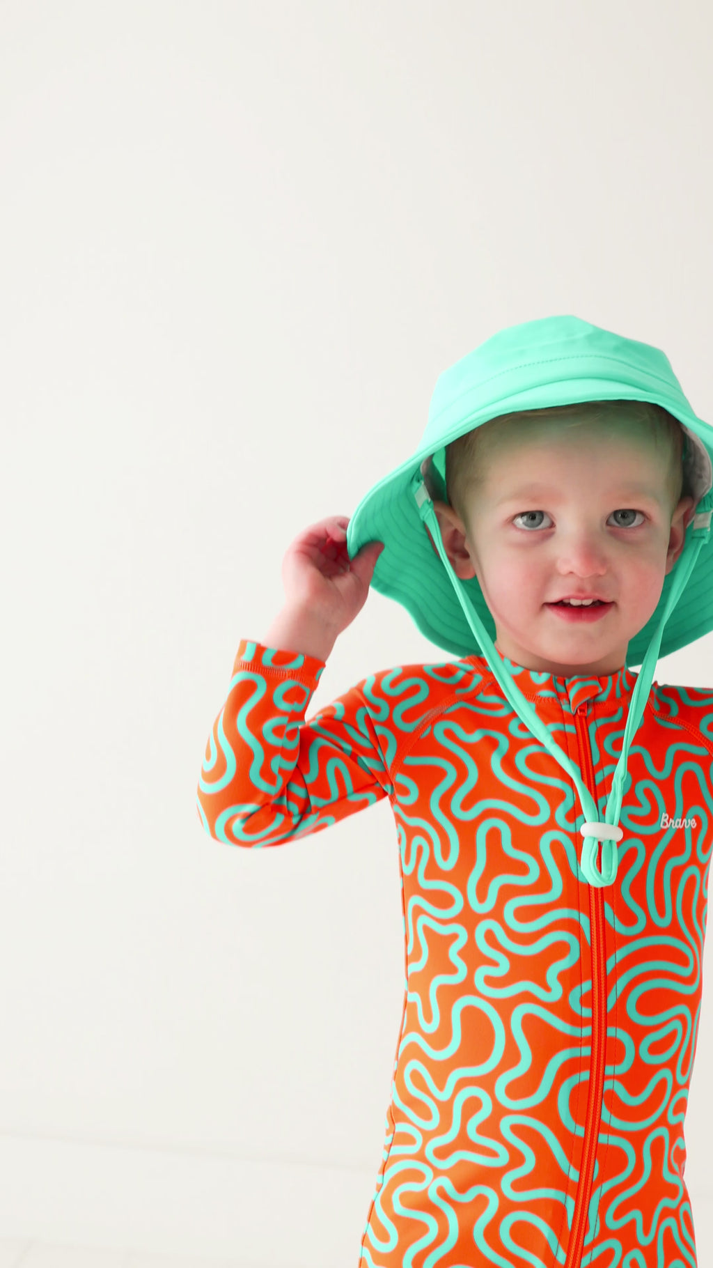 Child wearing a colorful boy swimsuit with a squiggly pattern and a turquoise bucket hat on a white background