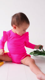 Baby wearing a bright pink one-piece swimsuit sitting on a white background with a yellow towel and green leaves.