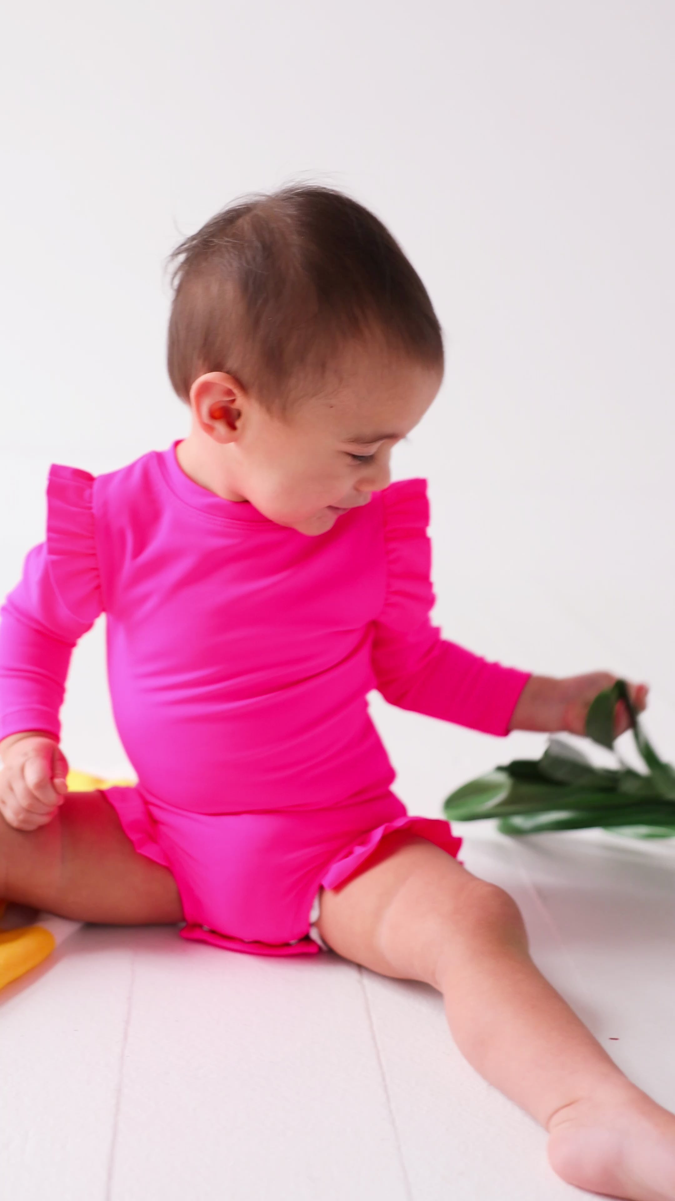 Baby wearing a bright pink one-piece swimsuit sitting on a white background with a yellow towel and green leaves.