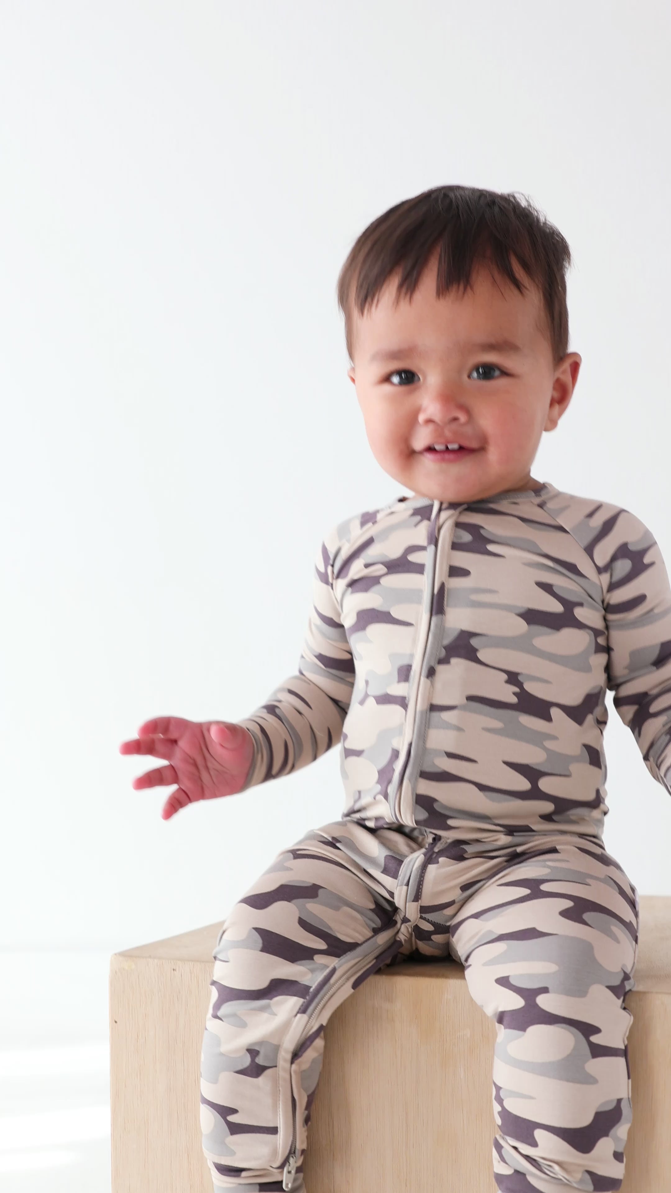 Video of a baby wearing a camouflage onesie sitting next on a wooden surface, on a white background
