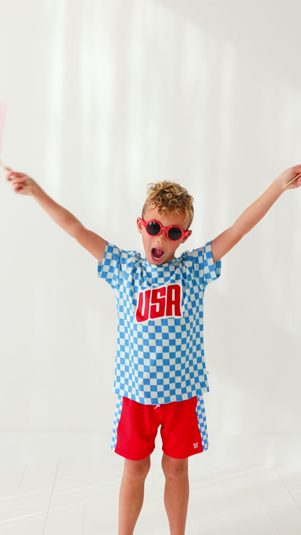 Child wearing a blue 'USA' t-shirt with checkered shorts, holding American flags on a white background