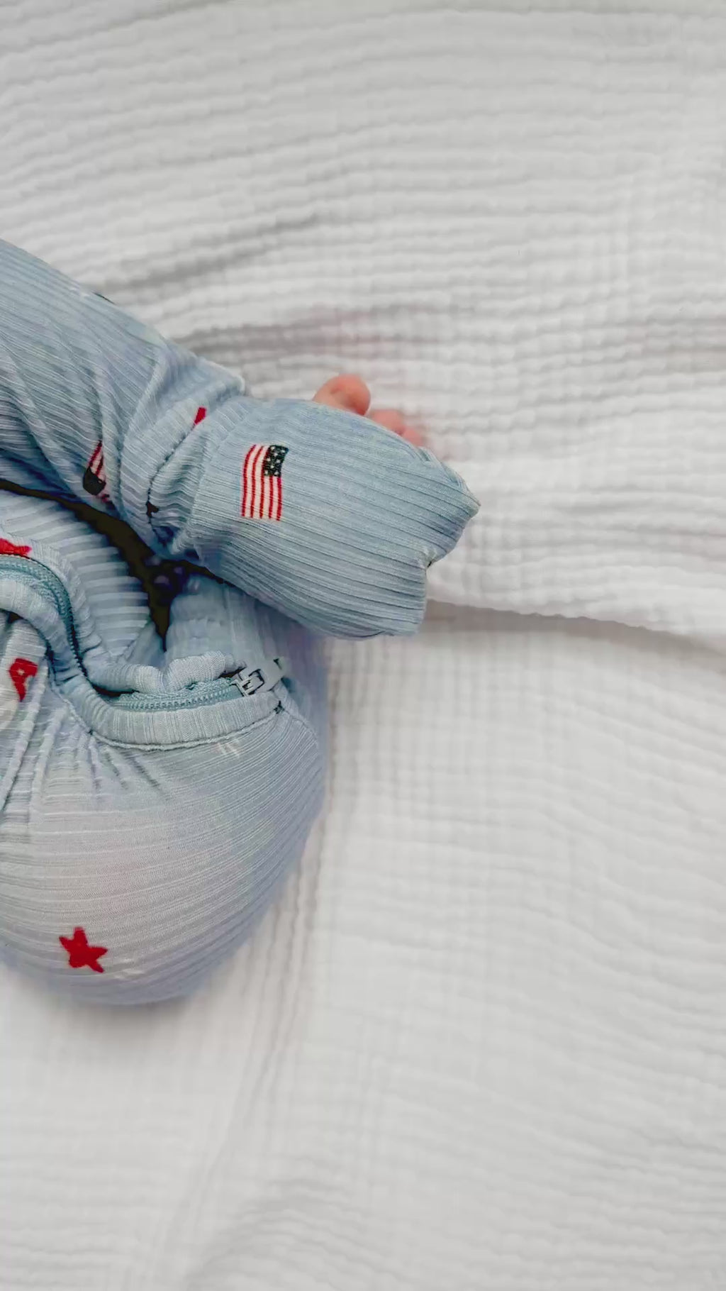 Video of a newborn baby sleeping in a blue onesie with stars and flags on a white textured background.