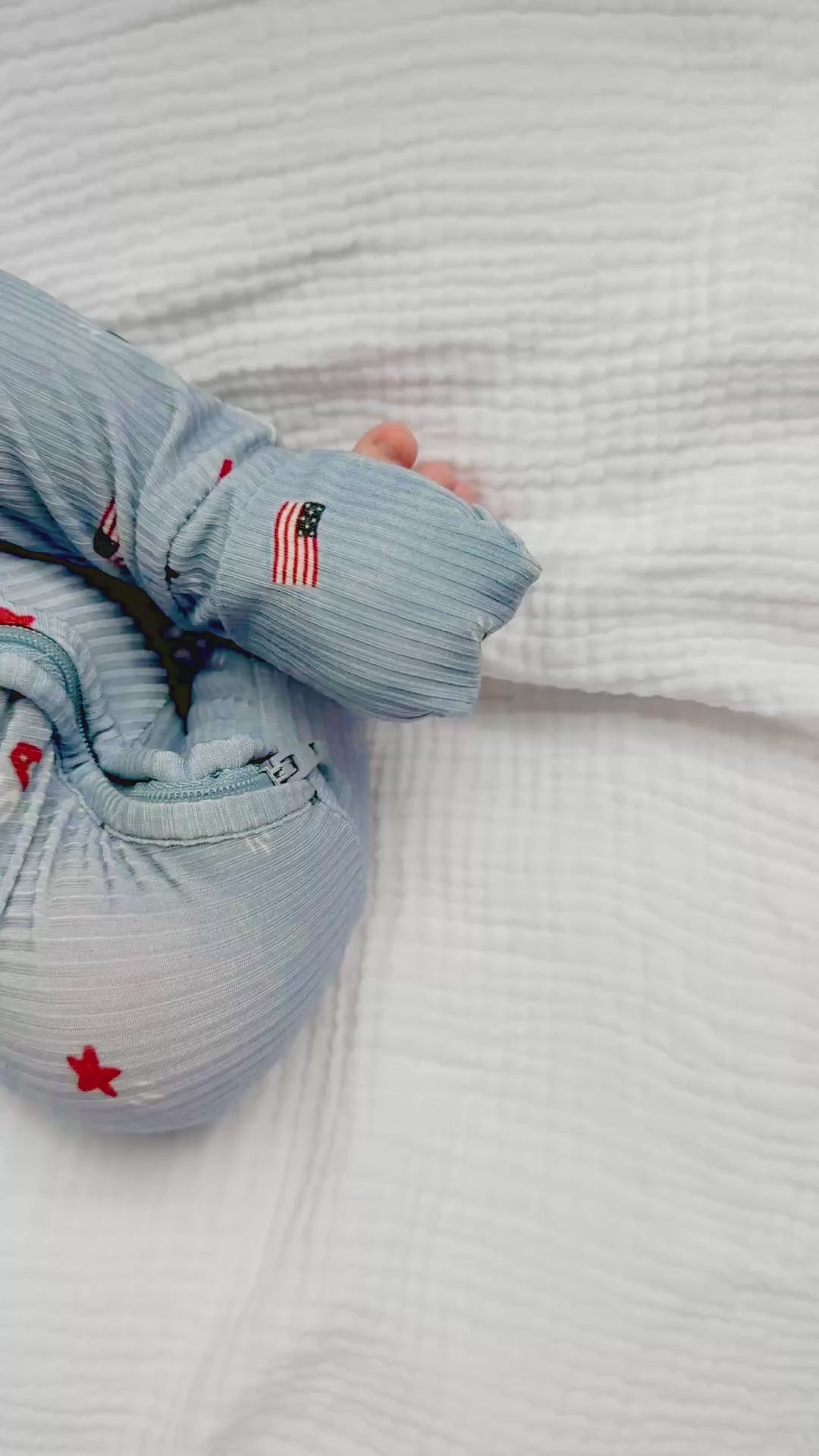 Video of a newborn baby sleeping in a blue onesie with stars and flags on a white textured background.