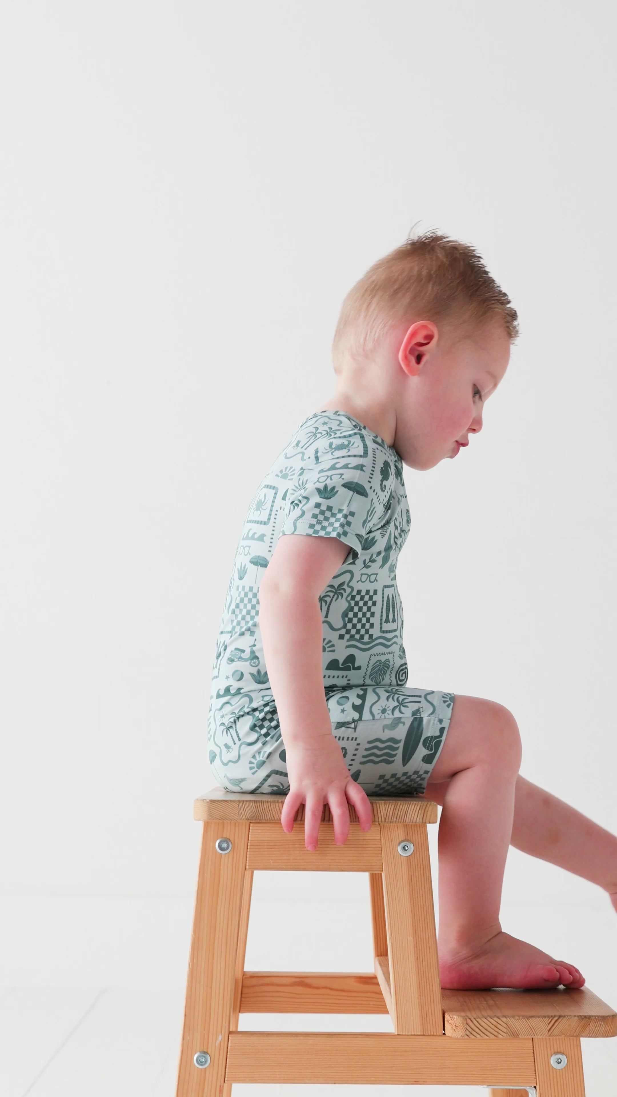 Child wearing a blue short romper with summer patterns sitting on a stool
