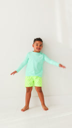 Child wearing neon yellow swim shorts with blue lightning bolt pattern and turquoise rash guard on a white background