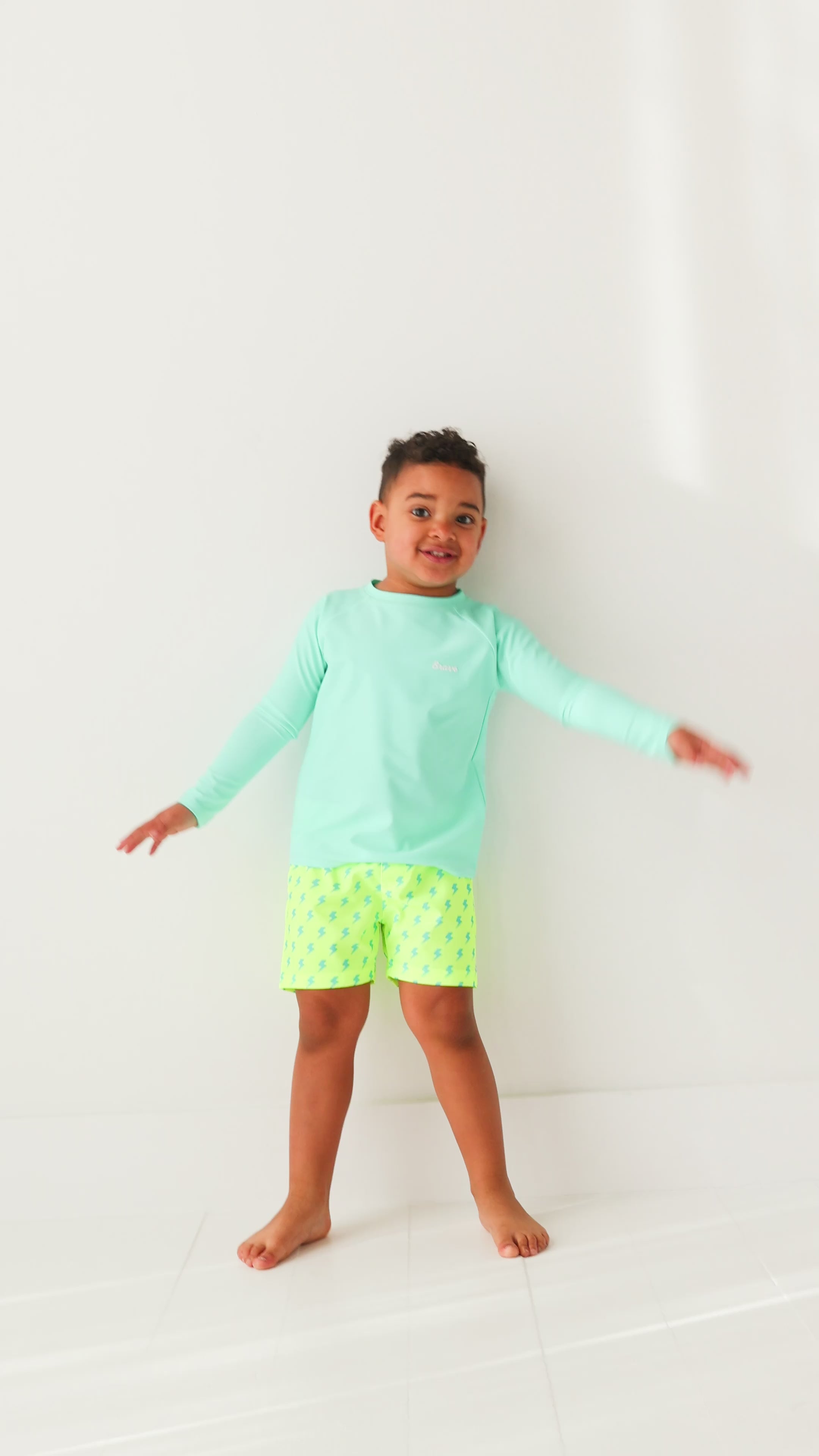 Child wearing neon yellow swim shorts with blue lightning bolt pattern and turquoise rash guard on a white background