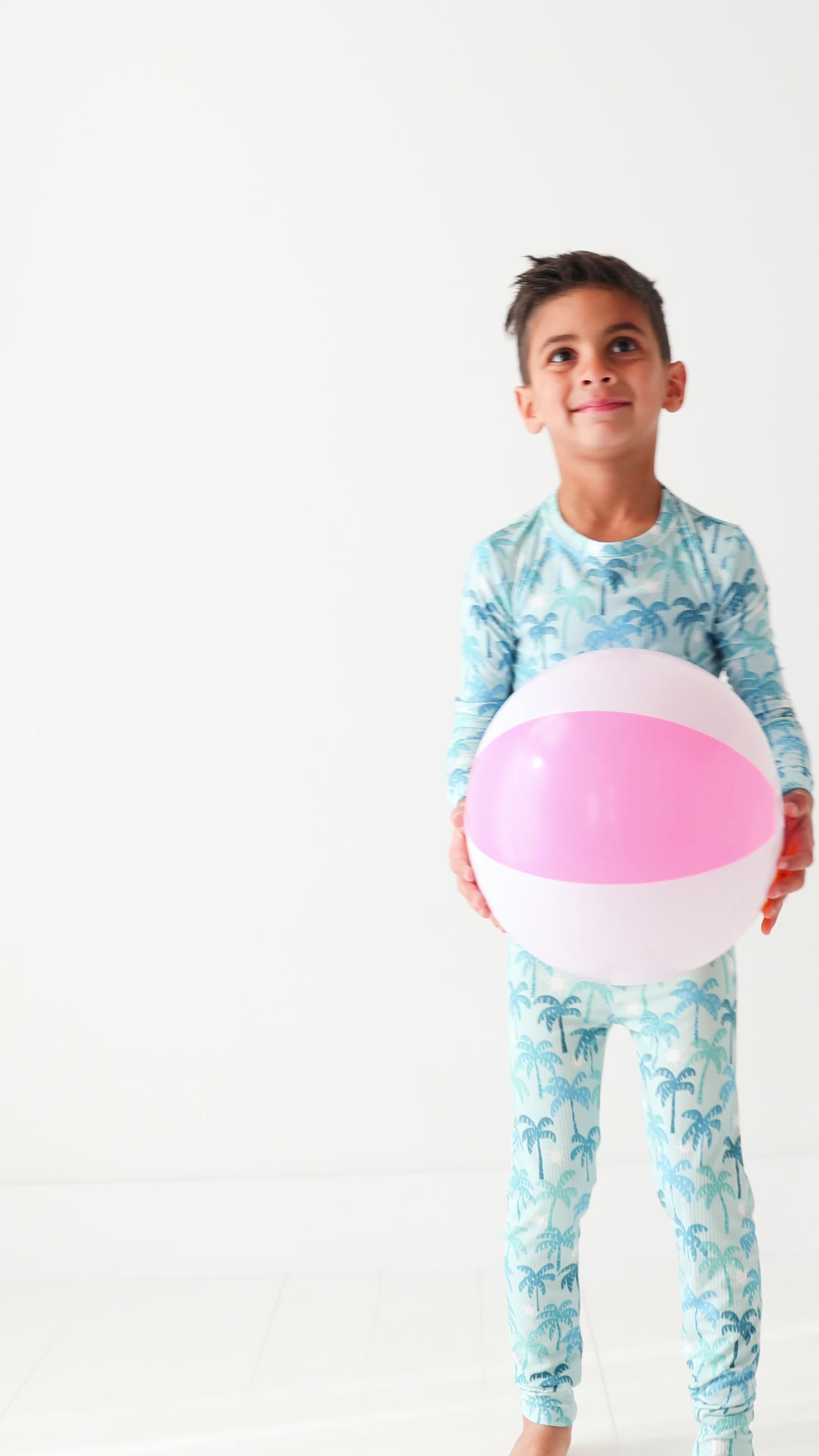 Child wearing a blue palm tree patterned pajama set holding a beach ball on a white background.