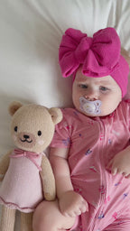 Video of a baby in pink shortie with teddy bear and pacifier, wearing a large pink bow headband.
