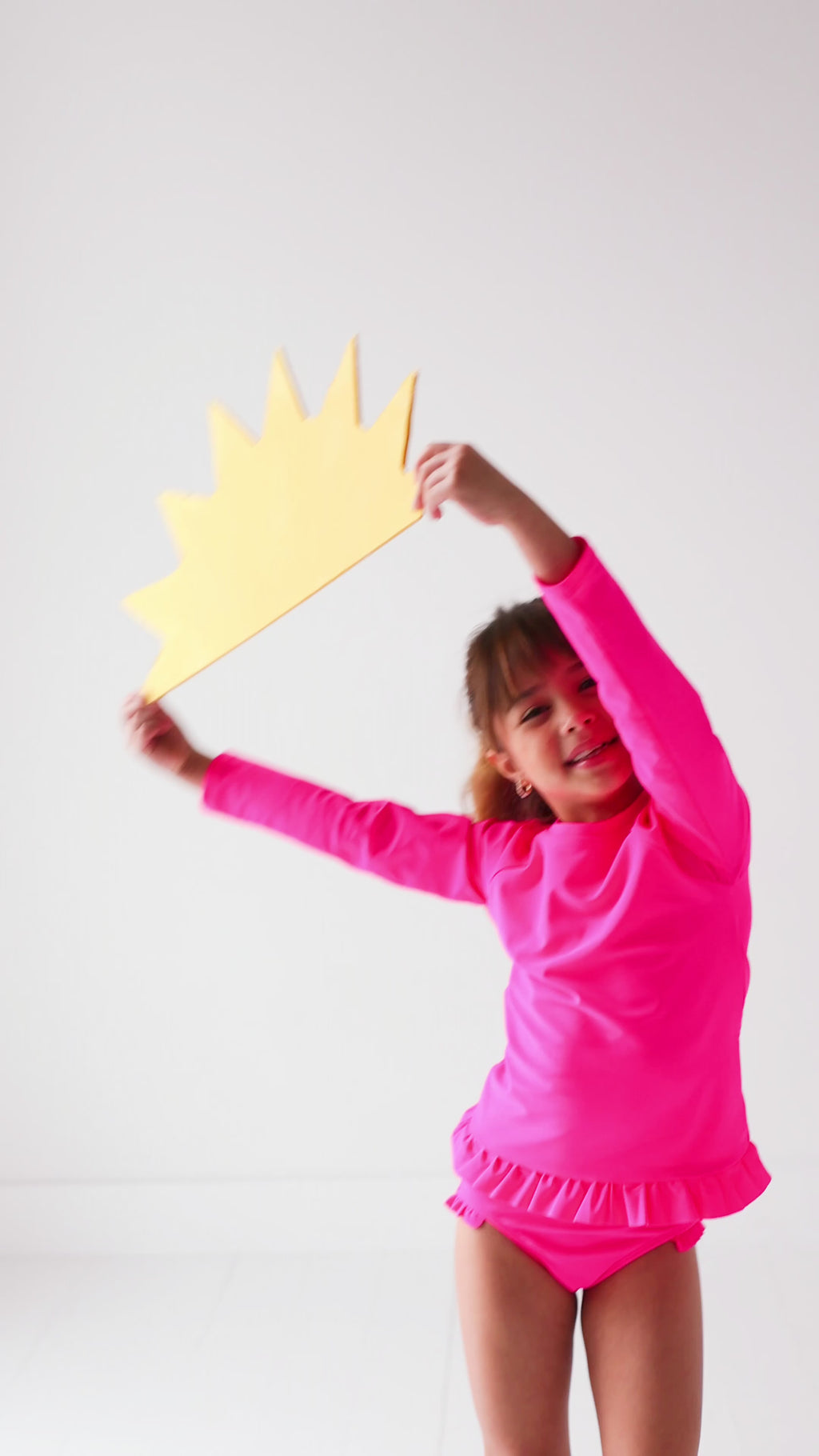 Child wearing a bright pink long sleeve two-piece swimsuit, holding a yellow sun-shaped object.
