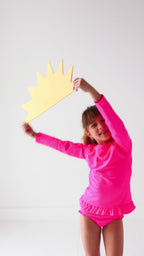 Child wearing a bright pink long sleeve two-piece swimsuit, holding a yellow sun-shaped object.