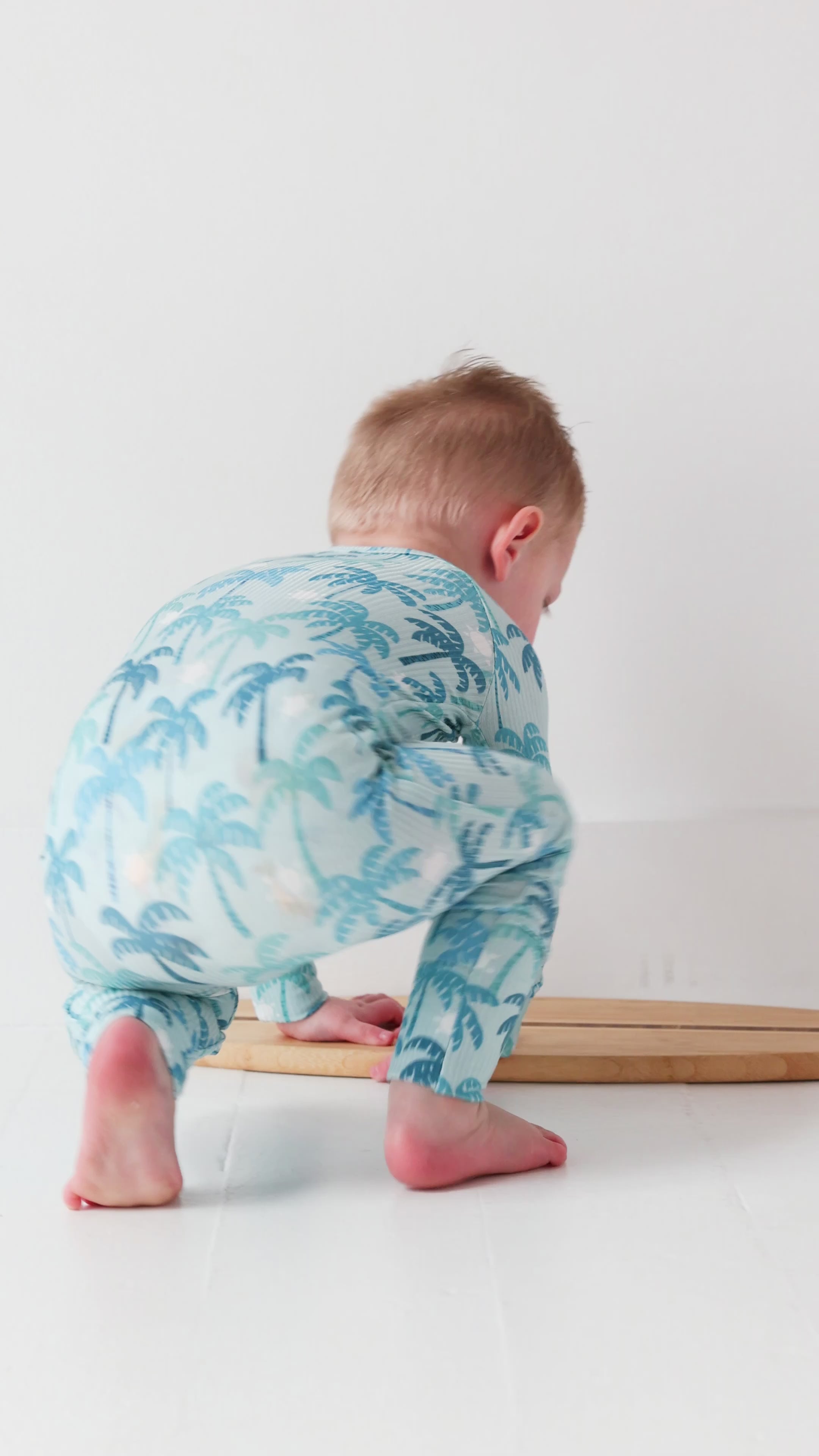 Child wearing a blue palm tree patterned onesie standing on a surfboard against a white background