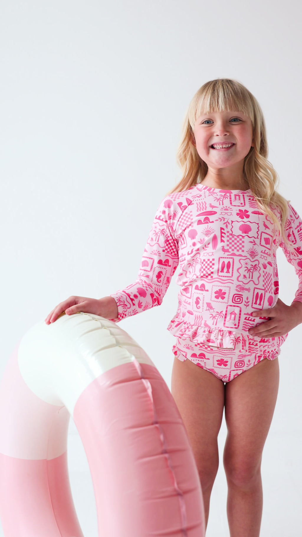 Child wearing a pink and white patterned long sleeve two-piece swimsuit on a white background holding a pink floatie