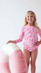 Child wearing a pink and white patterned long sleeve two-piece swimsuit on a white background holding a pink floatie