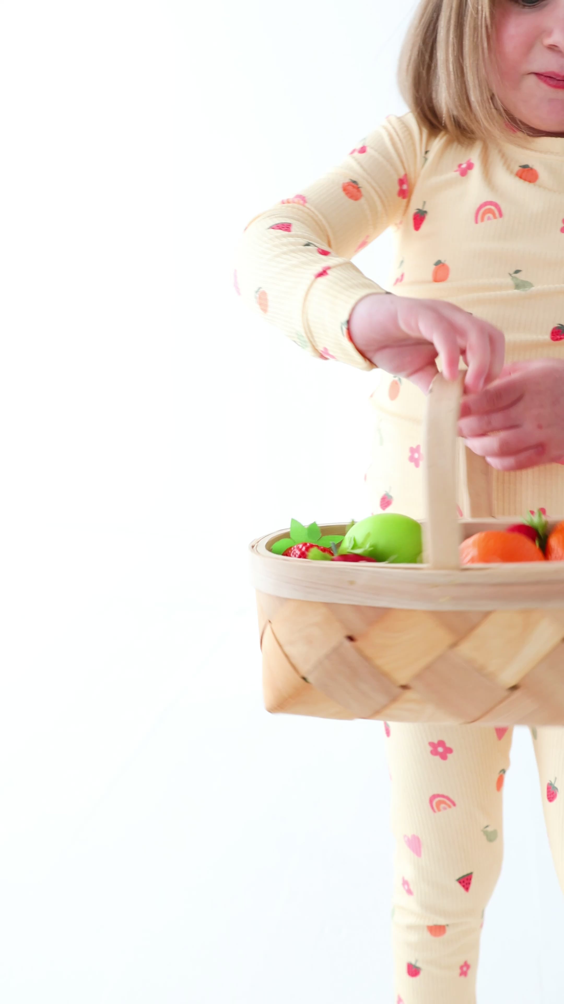 Child wearing a yellow pajama set with fruit pattern, holding a basket of fruits on a white background