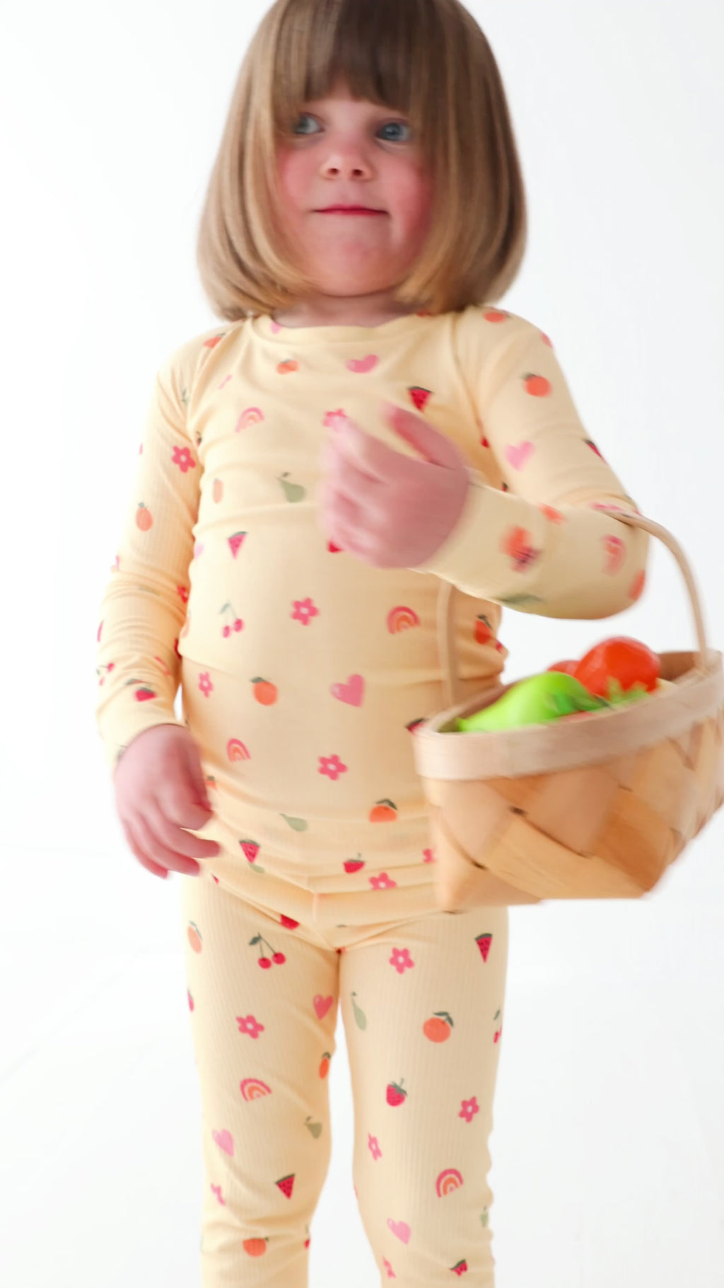 Child wearing a yellow pajama set with fruit pattern, holding a basket of fruits on a white background