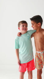 Child wearing orange and blue squiggly-patterned swim shorts and another child wearing a blue summer shirt and coral checkered swim shorts