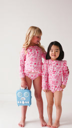 Two children wearing a pink and white patterned long sleeve one-piece and two-piece swimsuit holding hands with a blue radio toy.