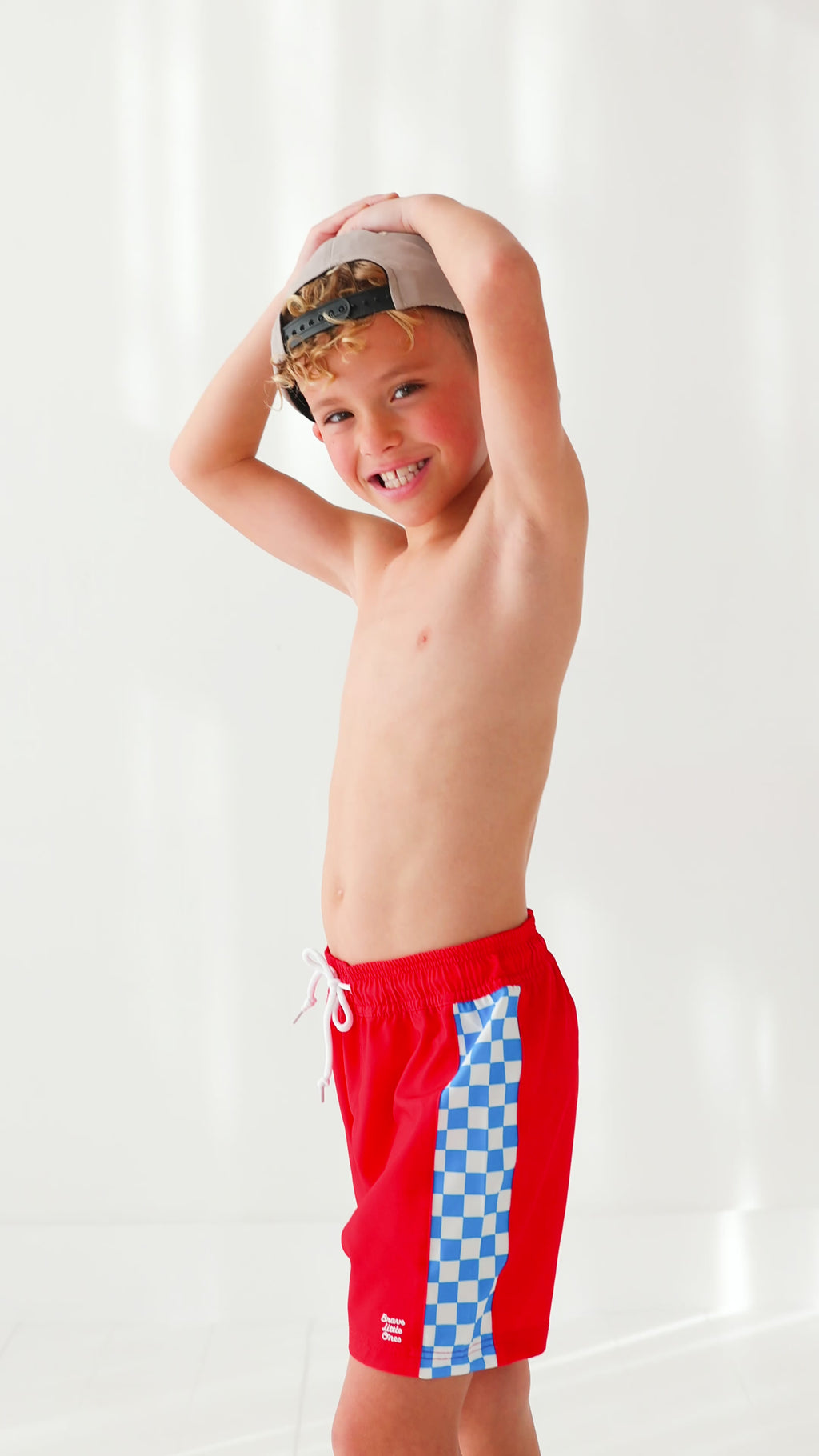 Young boy wearing red swim trunks and taupe hat on a white background