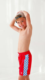 Young boy wearing red swim trunks and taupe hat on a white background