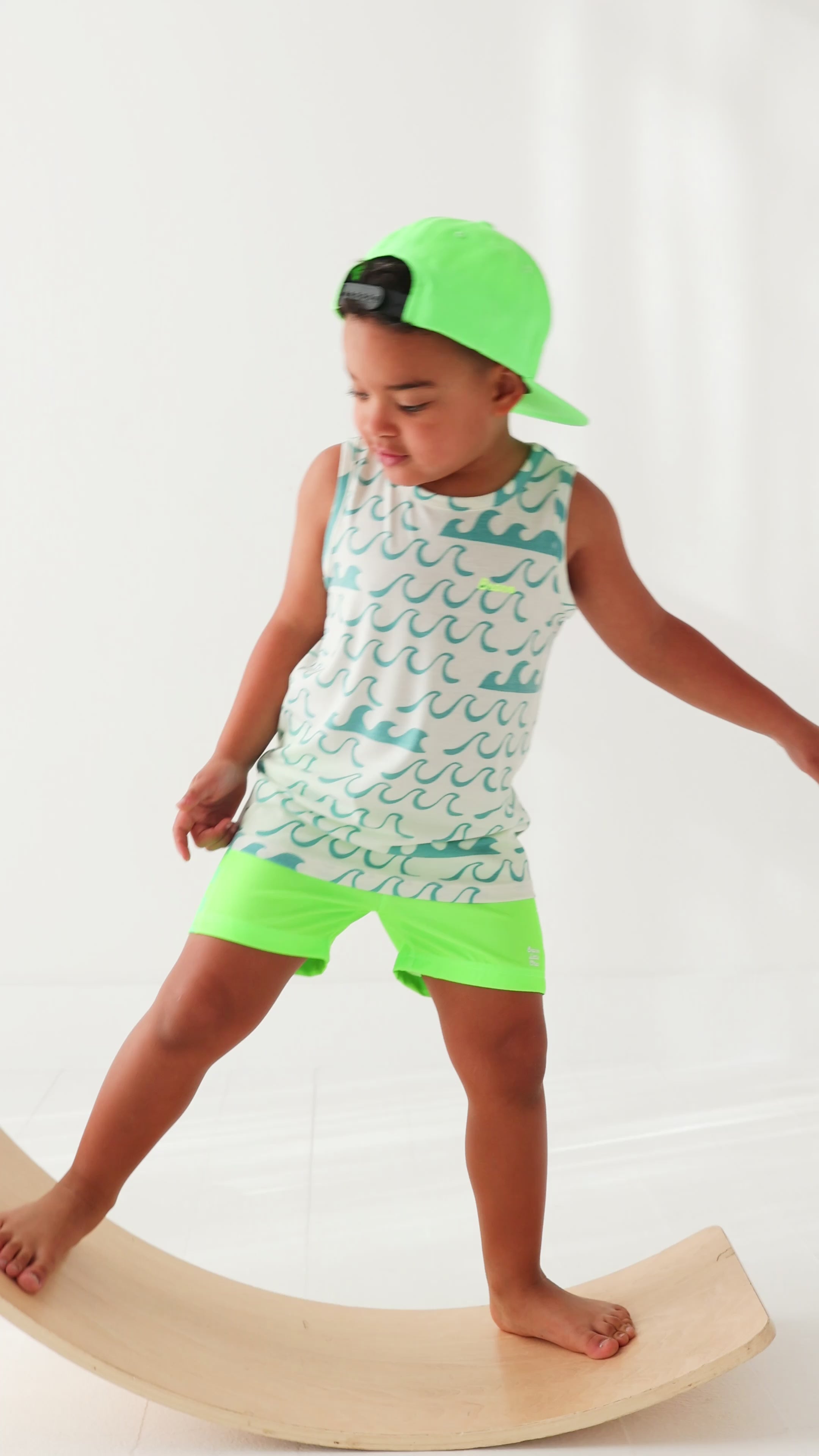 Child wearing a green checkered swim trunks, a wave-patterned top and a green trucker hat on a white background
