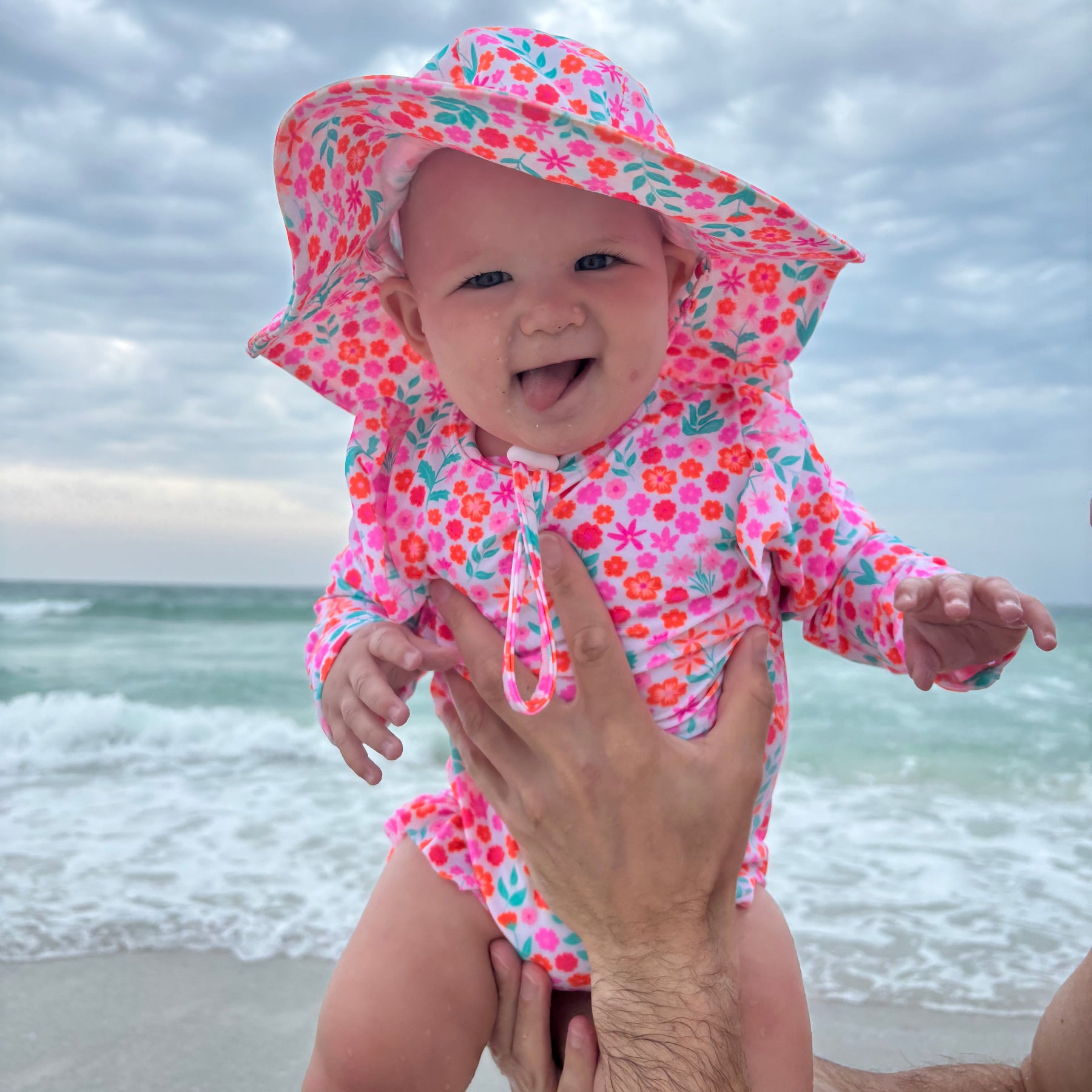 Baby in a colorful floral one-piece swimsuit and matching sun hat on a beach
