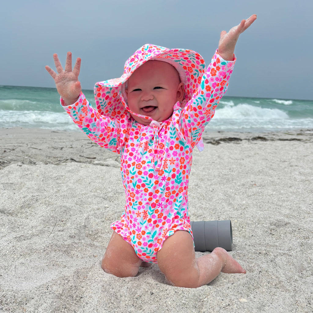 Baby in a colorful floral one-piece swimsuit and matching sun hat on a beach