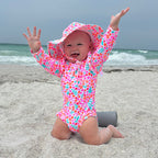 Baby in a colorful floral one-piece swimsuit and matching sun hat on a beach