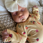 Baby wearing a yellow outfit with fruit patterns and a large white bow on head, lying on textured fabric.